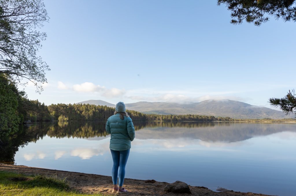 Person standing by Loch Garten at sunrise, overlooking calm water and forested hills
