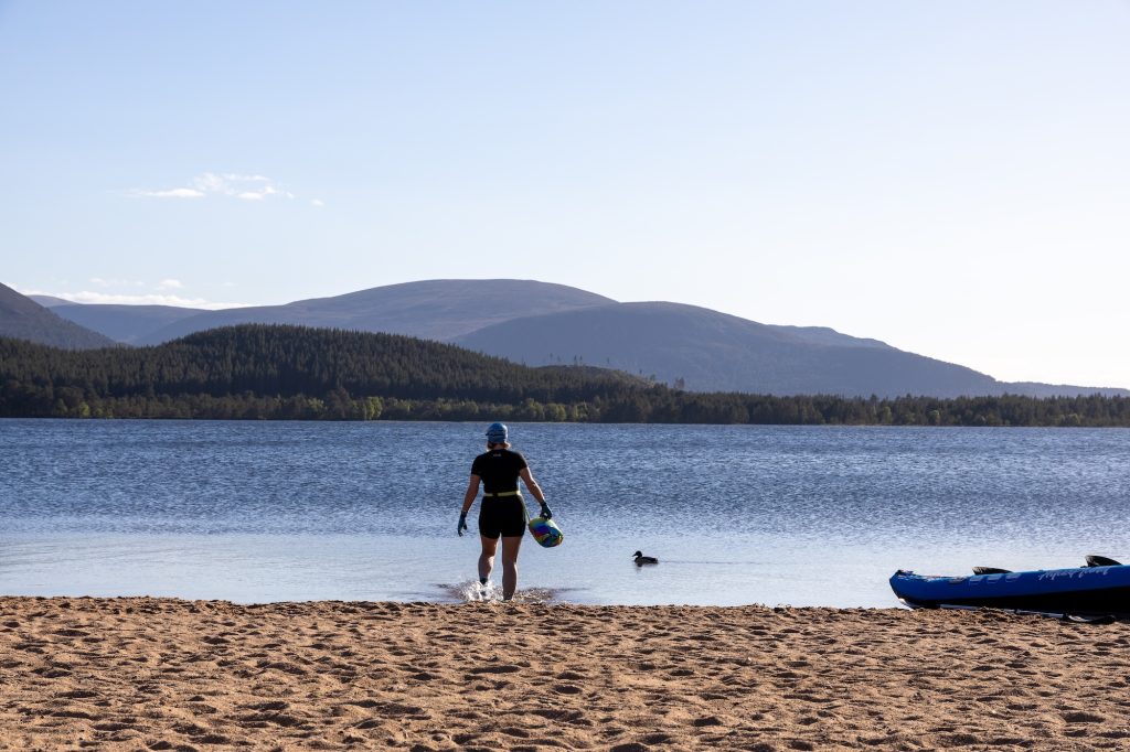 Person walking into Loch Morlich wild swimming on sandy beach, surrounded by Highland hills